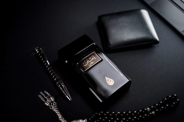 A dark-themed flat lay featuring a black perfume bottle with gold Arabic script and detailing, placed beside a black wallet. A sleek black and silver pen and a string of black prayer beads with a silver tassel complement the arrangement. The overall setup is positioned against a matte black background.