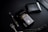 A dark-themed flat lay featuring a black perfume bottle with gold Arabic script and detailing, placed beside a black wallet. A sleek black and silver pen and a string of black prayer beads with a silver tassel complement the arrangement. The overall setup is positioned against a matte black background.