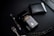 A dark-themed flat lay featuring a black perfume bottle with gold Arabic script and detailing, placed beside a black wallet. A sleek black and silver pen and a string of black prayer beads with a silver tassel complement the arrangement. The overall setup is positioned against a matte black background.