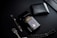 A dark-themed flat lay featuring a black perfume bottle with gold Arabic script and detailing, placed beside a black wallet. A sleek black and silver pen and a string of black prayer beads with a silver tassel complement the arrangement. The overall setup is positioned against a matte black background.