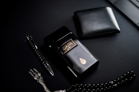 A dark-themed flat lay featuring a black perfume bottle with gold Arabic script and detailing, placed beside a black wallet. A sleek black and silver pen and a string of black prayer beads with a silver tassel complement the arrangement. The overall setup is positioned against a matte black background.