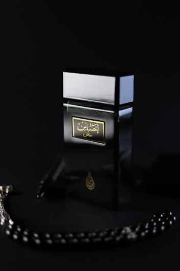 A black perfume bottle with Arabic writing on the label, placed against a dark background. The bottle has a sleek, modern design with a reflective surface. There appears to be a string of black beads with white accents partially visible next to the bottle.