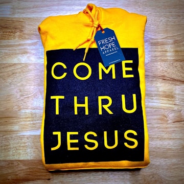 A folded yellow hoodie with a design reading 'COME THRU JESUS' in large, bold yellow letters on a black background. A blue tag attached to the hoodie reads 'FRESH HOPE APPAREL'. The hoodie is placed on a wooden surface.