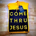 A folded yellow hoodie with a design reading 'COME THRU JESUS' in large, bold yellow letters on a black background. A blue tag attached to the hoodie reads 'FRESH HOPE APPAREL'. The hoodie is placed on a wooden surface.