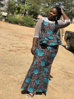 Woman wearing a flowing, patterned dress standing in a sunlit urban street.
