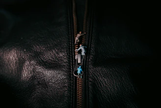 A close-up of a textured black surface with a zipper. Tiny figurines are depicted climbing on the zipper, creating a whimsical scene.