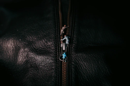 A close-up of a textured black surface with a zipper. Tiny figurines are depicted climbing on the zipper, creating a whimsical scene.