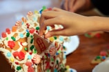 A hand decorates a gingerbread house with colorful candy pieces including red and white peppermint swirls, green and red gumdrops, and multicolored star and flower-shaped candies on a background of white icing.