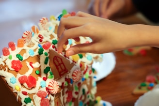 A hand decorates a gingerbread house with colorful candy pieces including red and white peppermint swirls, green and red gumdrops, and multicolored star and flower-shaped candies on a background of white icing.