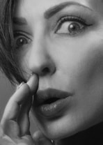 A close-up black and white photograph of a woman's face, capturing her wide, expressive eyes and slightly parted lips. Her hand touches her face near the mouth, emphasizing a dramatic and intense expression.