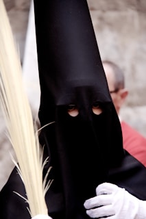 A person wearing a tall, black, pointed hood with eye holes, commonly associated with traditional religious processions. The individual is holding a palm frond and wearing white gloves.