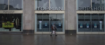 A storefront with large windows and closed metal security gates. The signs above the doors read 'TOPMAN' and 'TOPSHOP'. A sidewalk stretches across the bottom of the image with a person walking by, slightly blurred. Another sign in the window advertises a 50% sale. The scene suggests a quiet, possibly early or late time of day.
