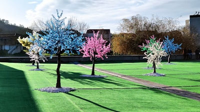 A series of artificial trees with bright, colorful lights are arranged in a landscaped area with green turf. Each tree is adorned with clusters of blue, pink, white, and yellow lights, creating a vibrant and festive atmosphere. The sky is mostly clear, and there are buildings and natural trees visible in the background.