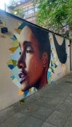A detailed mural on a wall depicting a realistic side profile of a person with geometric shapes and vivid colors in the background. The mural is situated on a pedestrian walkway, surrounded by greenery and urban elements.