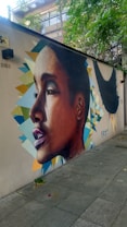 A detailed mural on a wall depicting a realistic side profile of a person with geometric shapes and vivid colors in the background. The mural is situated on a pedestrian walkway, surrounded by greenery and urban elements.