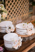 Three semi-naked cakes adorned with delicate peach and white flowers are beautifully arranged on circular wooden slabs. They rest on a rustic wooden table, partially surrounded by lush green foliage and a lattice wood backdrop. The floral arrangements add an elegant touch, with the soft tones contrasting against the natural wood textures.