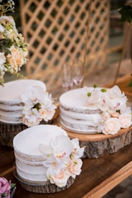 Three semi-naked cakes adorned with delicate peach and white flowers are beautifully arranged on circular wooden slabs. They rest on a rustic wooden table, partially surrounded by lush green foliage and a lattice wood backdrop. The floral arrangements add an elegant touch, with the soft tones contrasting against the natural wood textures.