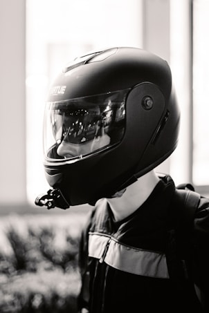 A monochromatic image featuring a person wearing a sleek, black motorcycle helmet with a clear visor. The helmet has branding on the side, and the person is dressed in a dark jacket. There's a blurred background with vertical elements.