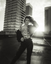 A person stands confidently on a rooftop with two tall buildings towering in the background. The image is in black and white, with sunlight creating a dramatic lens flare. The person is wearing a patterned top and black pants, with a jacket draped over one shoulder, and their hair blowing slightly in the wind.