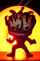 A stylized figure resembling a comic or movie character stands in front of a bright, circular orange light source. Its body is dark with a menacing expression and exaggerated features. The background and lighting create a dramatic and intense visual effect.