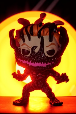 A stylized figure resembling a comic or movie character stands in front of a bright, circular orange light source. Its body is dark with a menacing expression and exaggerated features. The background and lighting create a dramatic and intense visual effect.