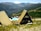 Two A-frame cabins with yellow roofs are situated in a mountainous landscape. A small group of people is sitting at picnic tables on a deck, enjoying the view. The background features a range of mountains with green and brown hues, and the foreground is filled with patches of grass and rocks.