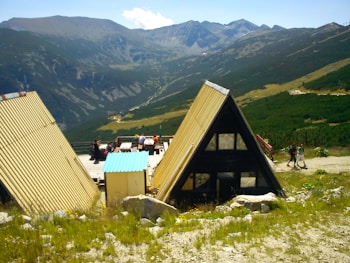 Two A-frame cabins with yellow roofs are situated in a mountainous landscape. A small group of people is sitting at picnic tables on a deck, enjoying the view. The background features a range of mountains with green and brown hues, and the foreground is filled with patches of grass and rocks.