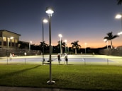 A tennis court under artificial lighting during dusk, with several people playing tennis. The surrounding area includes palm trees, a building with lit windows, and a well-maintained grassy area. Streetlights are also visible, illuminating the court and its surroundings.