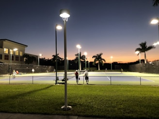 A tennis court under artificial lighting during dusk, with several people playing tennis. The surrounding area includes palm trees, a building with lit windows, and a well-maintained grassy area. Streetlights are also visible, illuminating the court and its surroundings.