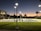 A tennis court under artificial lighting during dusk, with several people playing tennis. The surrounding area includes palm trees, a building with lit windows, and a well-maintained grassy area. Streetlights are also visible, illuminating the court and its surroundings.