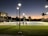 A tennis court under artificial lighting during dusk, with several people playing tennis. The surrounding area includes palm trees, a building with lit windows, and a well-maintained grassy area. Streetlights are also visible, illuminating the court and its surroundings.