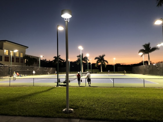 A tennis court under artificial lighting during dusk, with several people playing tennis. The surrounding area includes palm trees, a building with lit windows, and a well-maintained grassy area. Streetlights are also visible, illuminating the court and its surroundings.