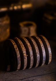 A rusted, large metal coil lies horizontally. The background is dark and out of focus, highlighting the corroded texture. The lighting is warm, casting dramatic shadows on the surface.