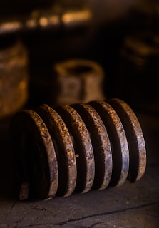 A rusted, large metal coil lies horizontally. The background is dark and out of focus, highlighting the corroded texture. The lighting is warm, casting dramatic shadows on the surface.