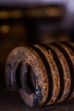 A detailed close-up of an aged and rusty coiled spring. The focus is on the intricate texture of the rust and the worn metal surface, showcasing signs of wear and corrosion. The background is blurred and dark, emphasizing the spring's details.