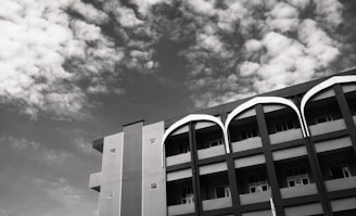 A four-story building with an architectural design featuring curved arches above its windows. The structure showcases a combination of vertical and horizontal lines, set against a partly cloudy sky backdrop. The photo is captured in black and white, creating a striking contrast between the building and the sky.