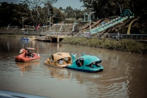 Colorful paddle boats in the shape of animals float on a calm body of water. In the background, a vibrant, multi-colored staircase and flags decorate the landscape, with trees lining the horizon. People are enjoying their time on the water, and there is a sense of leisure and playfulness.