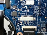 A detailed view of a blue printed circuit board with various electronic components such as capacitors, resistors, and integrated circuits. The board features visible traces and connections, along with labeled components and reference numbers.