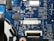 A detailed view of a blue printed circuit board with various electronic components such as capacitors, resistors, and integrated circuits. The board features visible traces and connections, along with labeled components and reference numbers.