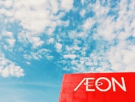 A clear blue sky with scattered white clouds provides a backdrop for a prominent red sign featuring the word 'AEON' in white letters.
