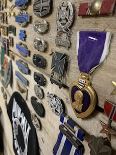A collection of military medals and badges displayed on a wooden board, including a prominent medal with a purple ribbon and gold border. Various other insignias and emblems in different shapes, sizes, and colors complement the main medal, indicating a display of honor and achievement.