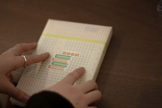 A pair of hands are wrapping or handling a book that is wrapped in patterned paper with a design featuring the word 'BOOK' and illustrations of books. The background is a wooden surface.