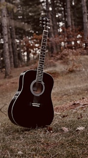 A black acoustic guitar is standing upright on a grassy forest floor surrounded by fallen leaves. The background features tall trees with some autumn foliage, indicating a natural, wooded setting.