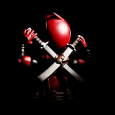 A stylized, action figure of a masked character holding two swords crossed behind its back. The figure is predominantly red and black with metallic accents on the swords. The lighting creates a dramatic contrast, highlighting the shiny texture of the character's suit against a dark background.