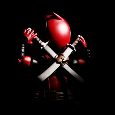 A stylized, action figure of a masked character holding two swords crossed behind its back. The figure is predominantly red and black with metallic accents on the swords. The lighting creates a dramatic contrast, highlighting the shiny texture of the character's suit against a dark background.