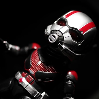 A toy or action figure of a superhero in a detailed metallic helmet with red and silver colors. The figure is dressed in a suit that combines red, black, and silver elements, set against a dark background.