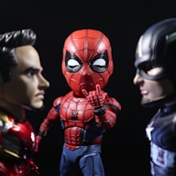 Three stylized superhero figures are arranged in a dramatic pose. They include a character in a red and black web-patterned suit standing between two other figures. The one on the left has metallic red and gold armor with a focused expression, while the one on the right wears a blue and silver suit featuring a prominent star, appearing determined.