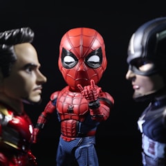 Three stylized superhero figures are arranged in a dramatic pose. They include a character in a red and black web-patterned suit standing between two other figures. The one on the left has metallic red and gold armor with a focused expression, while the one on the right wears a blue and silver suit featuring a prominent star, appearing determined.