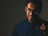 A young man wearing glasses and a dark shirt is depicted against a dark background. His facial expression suggests confusion or frustration, and he appears to be gesturing as if talking on the phone with his hand formed into a phone shape.