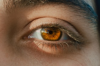 A close-up of a human eye with striking amber coloration and intricate patterns visible in the iris. The surrounding skin is textured, with a few visible eyelashes casting shadows.